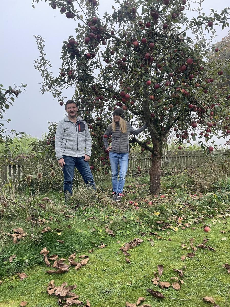 Gerhard und Martina Sedlmeier stehen in ihrem herbstlichen Garten unter einem mit Früchten behangenen Apfelbaum. Sie lächeln in die Kamera, umgeben von grüner Natur und einem vernebelten Feld im Hintergrund.