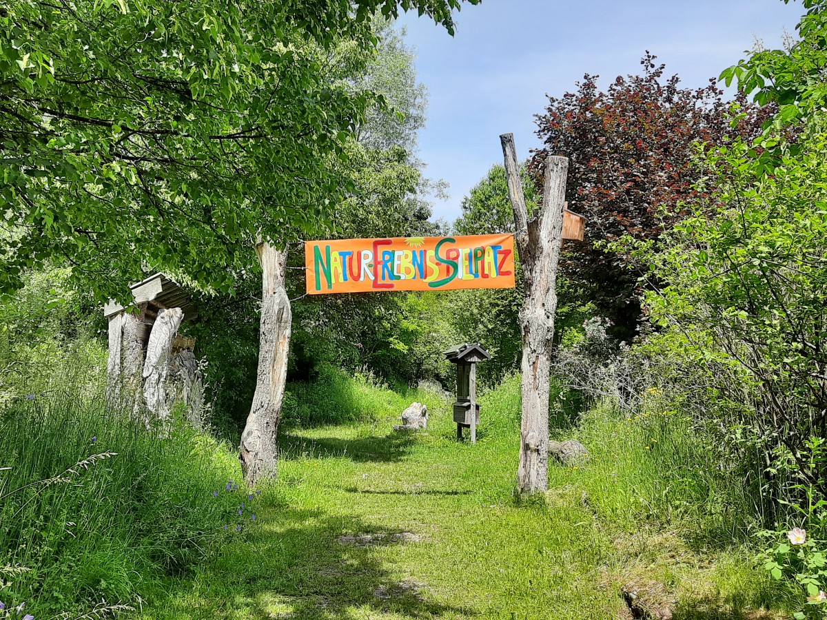 Zwei Holzstämme halten das Schild des Natur-Erlebnis-Spielplatzes Eglfing und markieren so den Eingang.